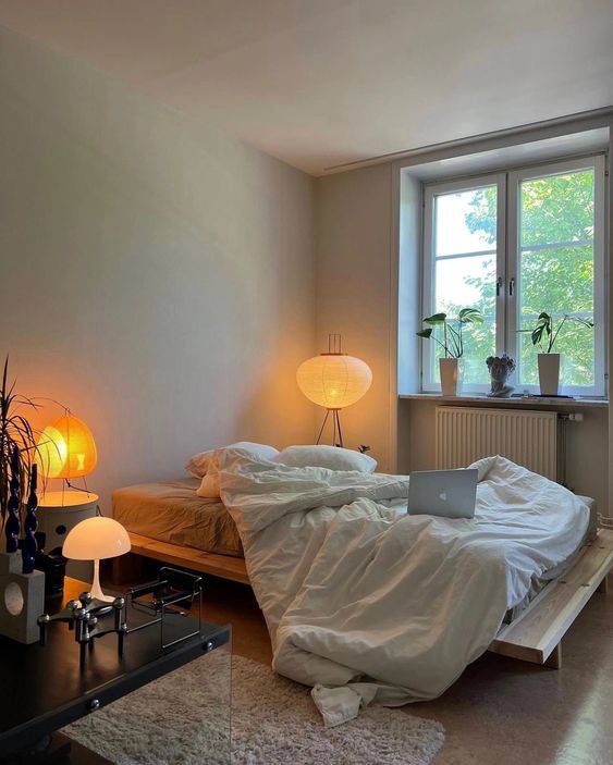 Minimalist cosy bedroom in white, beige, and grey with an unmade bed, soft lamp lighting, and an open laptop on the nightstand, creating a cozy work atmosphere.